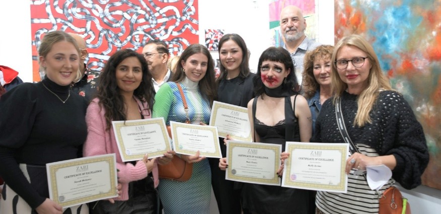 Six female students holding certificates stand in front of a wall of artwork.
